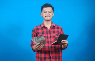 Kırmızı kareli gömlek giyen Asyalı adam tasarruf defteri ve dolar banknotlarını mavi arka planda izole ediyor. İş ve finans kavramı