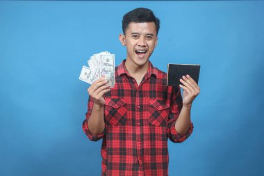 Kırmızı kareli gömlek giyen Asyalı adam tasarruf defteri ve dolar banknotlarını mavi arka planda izole ediyor. İş ve finans kavramı