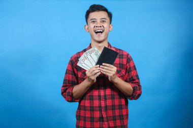 Kırmızı kareli gömlek giyen Asyalı adam tasarruf defteri ve dolar banknotlarını mavi arka planda izole ediyor. İş ve finans kavramı