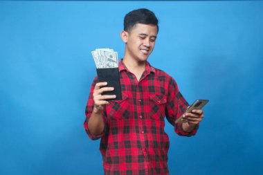 Genç adam elinde para ve akıllı telefon dolu bir tasarruf kitabı tutarken gülümsüyor. Finansal başarısını ve internet bankacılığını kutluyor..