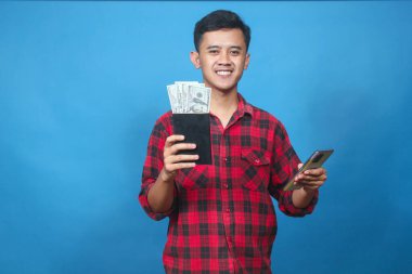 Genç adam elinde para ve akıllı telefon dolu bir tasarruf kitabı tutarken gülümsüyor. Finansal başarısını ve internet bankacılığını kutluyor..