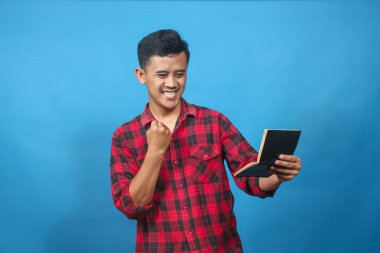 Genç adam başarısını mavi bir arka planda mutlu bir şekilde kutlarken gülümseyerek tasarruf defterini hevesle okuyor..