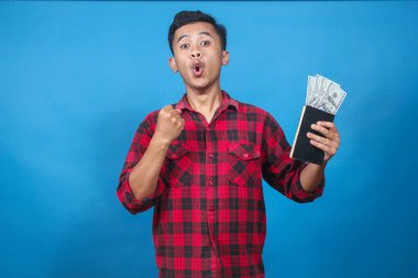 Şok olmuş genç bir adam elinde para dolu bir tasarruf defteri tutuyor ve yumruklarını mavi bir arka planda sıkıyor..