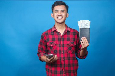 Genç adam elinde para ve akıllı telefon dolu bir tasarruf kitabı tutarken gülümsüyor. Finansal başarısını ve internet bankacılığını kutluyor..