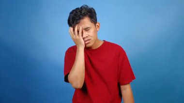 Bir tarafta baş ağrısı olan Asyalı bir adam mavi arka planda solgun bir ifade sergiliyor.