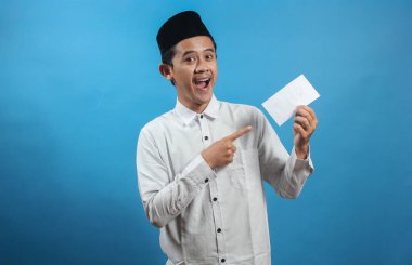 Asyalı Müslüman adam mavi arka planda mutlu bir şekilde izole edilmiş THR (tatil parası) zarfını işaret ediyor