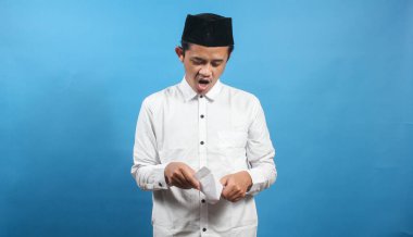 Beyaz gömlek giyen Asyalı Müslüman bir adam ve mavi arka planda dikkatlice izole edilmiş geleneksel Müslüman şapka açma zarfı.