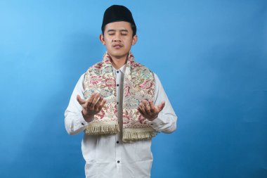 Boynunda beyaz gömlek, peci ve dua minderi olan Asyalı bir adam mavi arka planda ciddi ciddi dua ediyor.