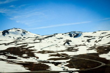 Güneşli bir günde, Jotunheimen Ulusal Parkı 'ndan geçen yüksek irtifalı ve dolambaçlı bir yol karla kaplı bir dağ arazisiyle çevrilidir..