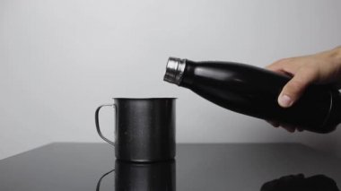 Erkek eli, siyah cam masadaki paslanmaz termo şişeden çelik bardağa su dökerken yakın plan. Beyaz rengin arkaplanı.