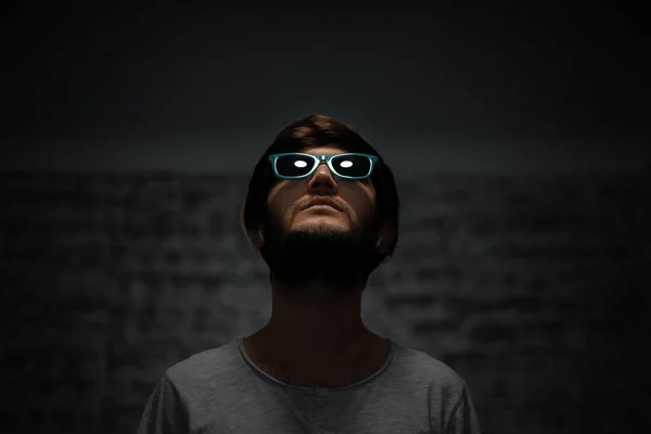 Dark portrait of young guy looking up, wearing sunglasses, using wireless earphones.