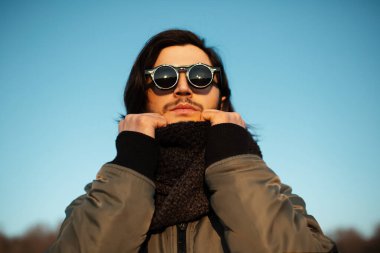 Güneş gözlüğü ve eşarp takan uzun saçlı genç adamın yakın plan portresi. Arka planda mavi gökyüzü.