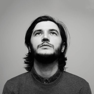 Gri arkaplana bakan uzun saçlı bir gencin siyah beyaz portresi.