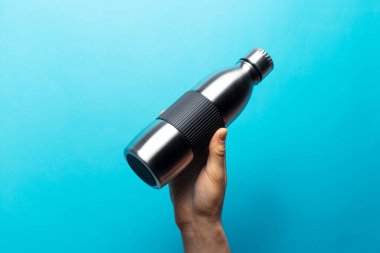 Mavi arka planda tekrar kullanılabilir çelik su şişesini tutan erkek eli yakın plan..