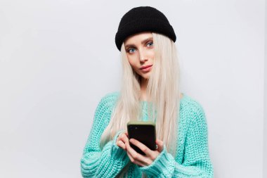 Beyaz arka planda akıllı telefon kullanan genç kızın stüdyo portresi. Siyah şapkalı ve mavi kazaklı kadın..