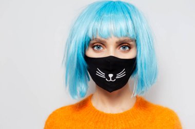 Coronavirus ve covid-19 'a karşı siyah tıbbi maske takan genç kızın yakın plan portresi. Beyaz stüdyonun portresi. Mavi saçlı, turuncu kazaklı bir kadın..