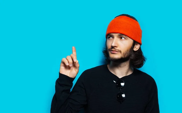 Studio portrait of young man pointing away against blue background. Wearing orange beanie hat and black sweater.
