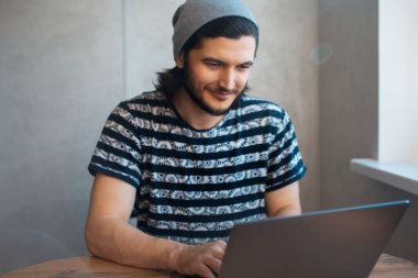 Pencerenin yanındaki laptopta çalışan neşeli bir gencin bulanık portresi..