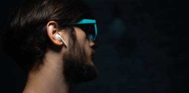 Kablosuz kulaklık kullanan, siyah arka planda mavi güneş gözlüğü takan genç bir adamın yakın plan portresi..