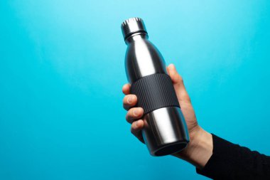 Çelik, yeniden kullanılabilir, kapak odaklı termo su şişesini mavi arka planda tutan erkek eli yakın plan..