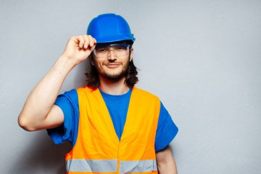 Güvenlik ekipmanı takan genç inşaat mühendisinin stüdyo portresi; mavi başlık, şeffaf gözlükler ve arka plandaki gri dokulu duvarda fotokopi alanı olan turuncu yelek.