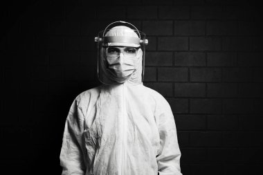 Koronavirüs ve covid-19 'a karşı PPE kıyafeti giyen kendine güvenen genç bir doktorun siyah beyaz portresi tuğla duvarın arkasında..