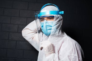 Koronavirüs ve kovid 19 'a karşı PPE kıyafeti giyen genç doktorun yakın plan portresi. Siyah tuğla duvarın arka planı. Güvenlik yüz kalkanını ellerimle tutuyorum..