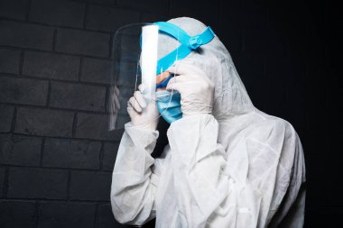 Koronavirüs ve kovid 19 'a karşı PPE kıyafeti giyen genç doktorun yakın plan portresi. Siyah tuğla duvarın arka planı. Güvenlik gözlüğü takıyorum..