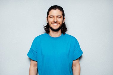 Gri desenli duvarın arka planında mavi gömlekli gülümseyen genç adam portresi.