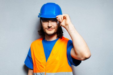 Güvenlik ekipmanı takan inşaat mühendisinin stüdyo portresi; mavi başlık ve gömlek, şeffaf gözlük ve gri desenli duvarın arka planında turuncu yelek.