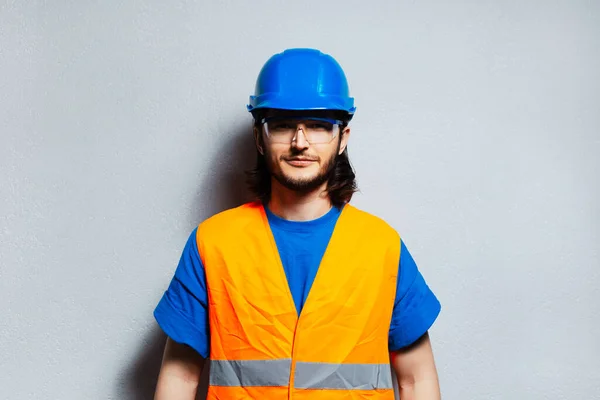 安全装置を身に着けている若い建設労働者のエンジニアの肖像 灰色のテクスチャ壁の背景に青のハード帽子 透明ゴーグルとオレンジのベスト — ストック写真
