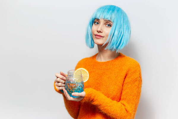 Studio portrait of young pretty girl with blue bob hairstyle, wearing orange sweater, holding glass mug with juice and piece of lemon. Background of white color.