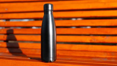 Fotokopi alanı olan turuncu ahşap bankın arka planında, termodinamik siyah bir şişe yakın plan. Yeniden kullanılabilir şişeler; sıfır atık; eko kavramı.