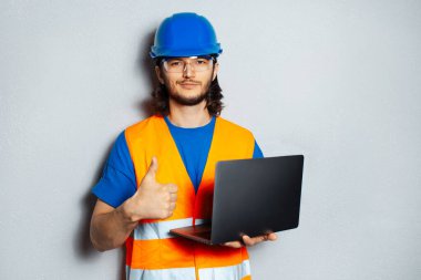 Kendine güvenen genç bir adamın portresi, güvenlik ekipmanı takan inşaat mühendisi, dizüstü bilgisayarı tutan ve arka planda gri desenli duvarda baş parmağını gösteren.