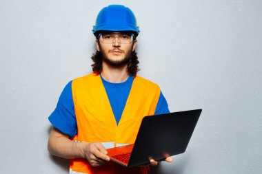 Kendine güveni olan genç bir adamın portresi, inşaat mühendisi güvenlik ekipmanı takıyor, gri arka planda dizüstü bilgisayar kullanıyor..