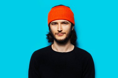 Mavi arka planda, turuncu şapkalı ve siyah kazaklı bir gencin stüdyo portresi..