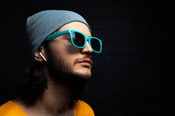 Studio portrait of young serious man with wireless earphone in ear, wearing sunglasses and hat on black background.
