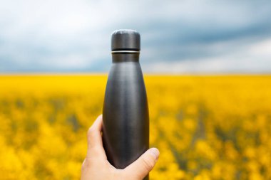 Arka planda siyah çelik termodinamik su şişesi tutan erkek eli, bulanık kolza sahip..