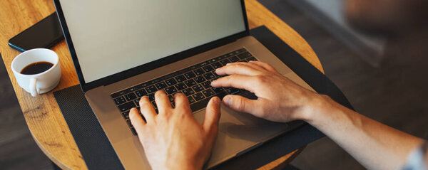 Close-up of male hands typing on laptop on wooden table near coffee cup and smartphone; panoramic banner, top view.