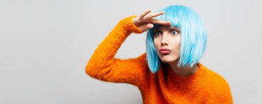 Stüdyo portresi, beyaz arka planda, alnında ellerle başka tarafa bakan güzel bir kızın resmi. Mavi peruk ve turuncu kazak giyiyor. Panoramik pankart görünümü.