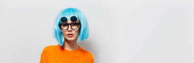 Stüdyoda mavi saçlı, siyah güneş gözlüğü takan ve beyaz arka planda turuncu süveter giyen hoş bir kızın portresi. Metin için kopyalama alanı olan panoramik afiş fotoğrafı.