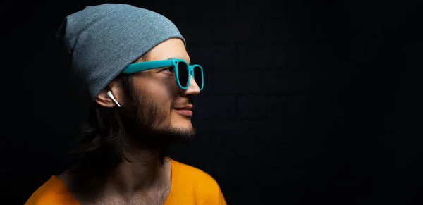 Close-up portrait of young smiling man with wireless earphone in ear on black background; wearing blue sunglasses. Panoramic banner view with copy space.