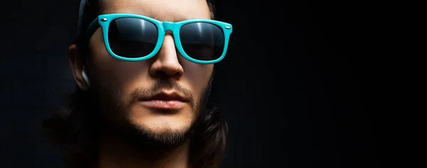 Close-up portrait of young serious man with wireless earphone in ear on black background; wearing blue sunglasses.