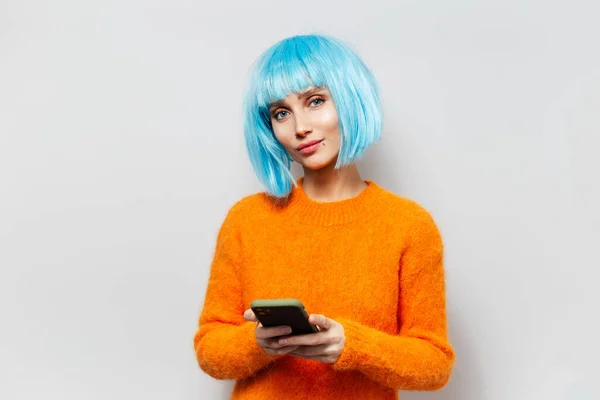 照片上的年轻漂亮姑娘手握智能手机 蓝色头发 白色背景上穿着橙色毛衣 — 图库照片