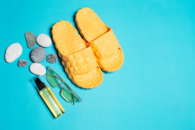 Top view of yellow slippers, sunglasses and oil tube near pebbles on blue background.