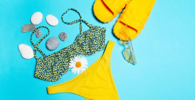 Close-up of green swimsuit, yellow sunglasses and slippers decorated with pebbles and chamomile flower on blue background.
