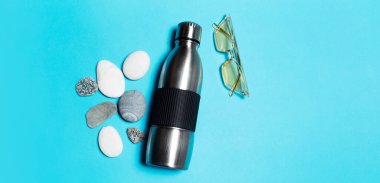 Top view of reusable steel thermo water bottle near yellow sunglasses and stones on blue background.