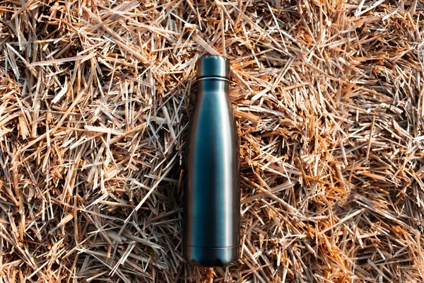Reusable steel thermo water bottle on background of dry haystack.
