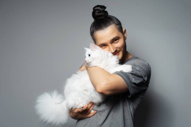 Gri arka planda beyaz kedisi olan genç bir adamın stüdyo portresi. Mavi gözlü evcil hayvan.