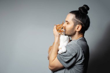 Stüdyo portresi. Kucağında kırmızı kedi olan genç adam. Gri arka planda evcil hayvanı öperken..